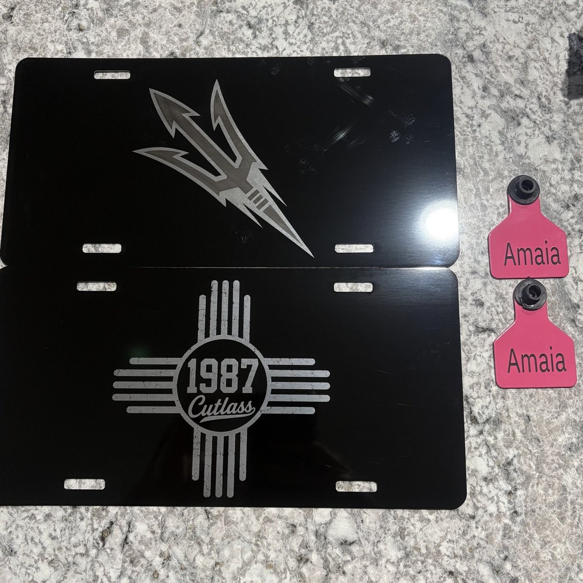 Two black engraved license plates beside pink personalized cattle ear tags, by KB Custom Creations